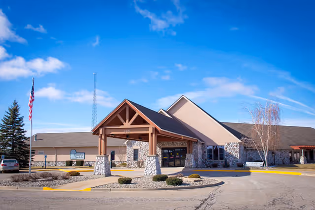 Exterior view of Country Gardens Assisted Living facility showing a single-story building with stone and beige siding, a covered entrance with wooden beams, a flagpole with an American flag, and a clear blue sky.
