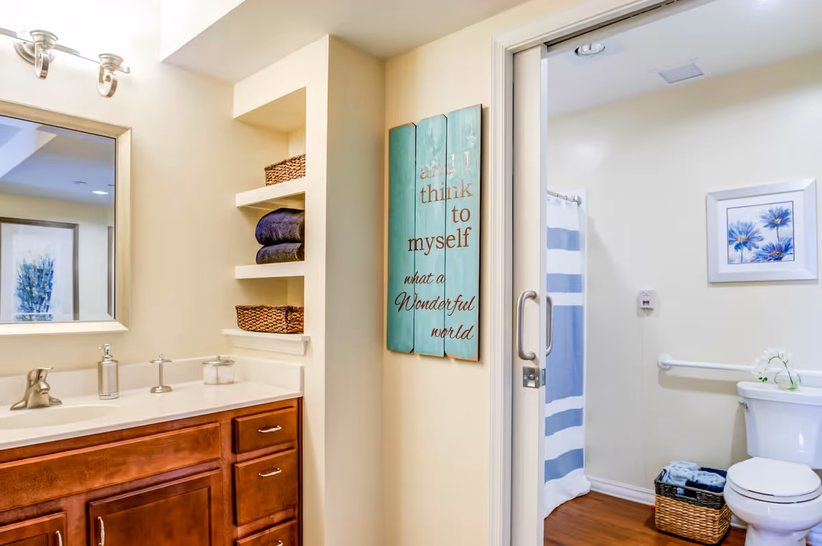 Bright bathroom with a wooden vanity and mirror, built-in shelves with towels and baskets, and a shower and toilet area with a striped curtain.
