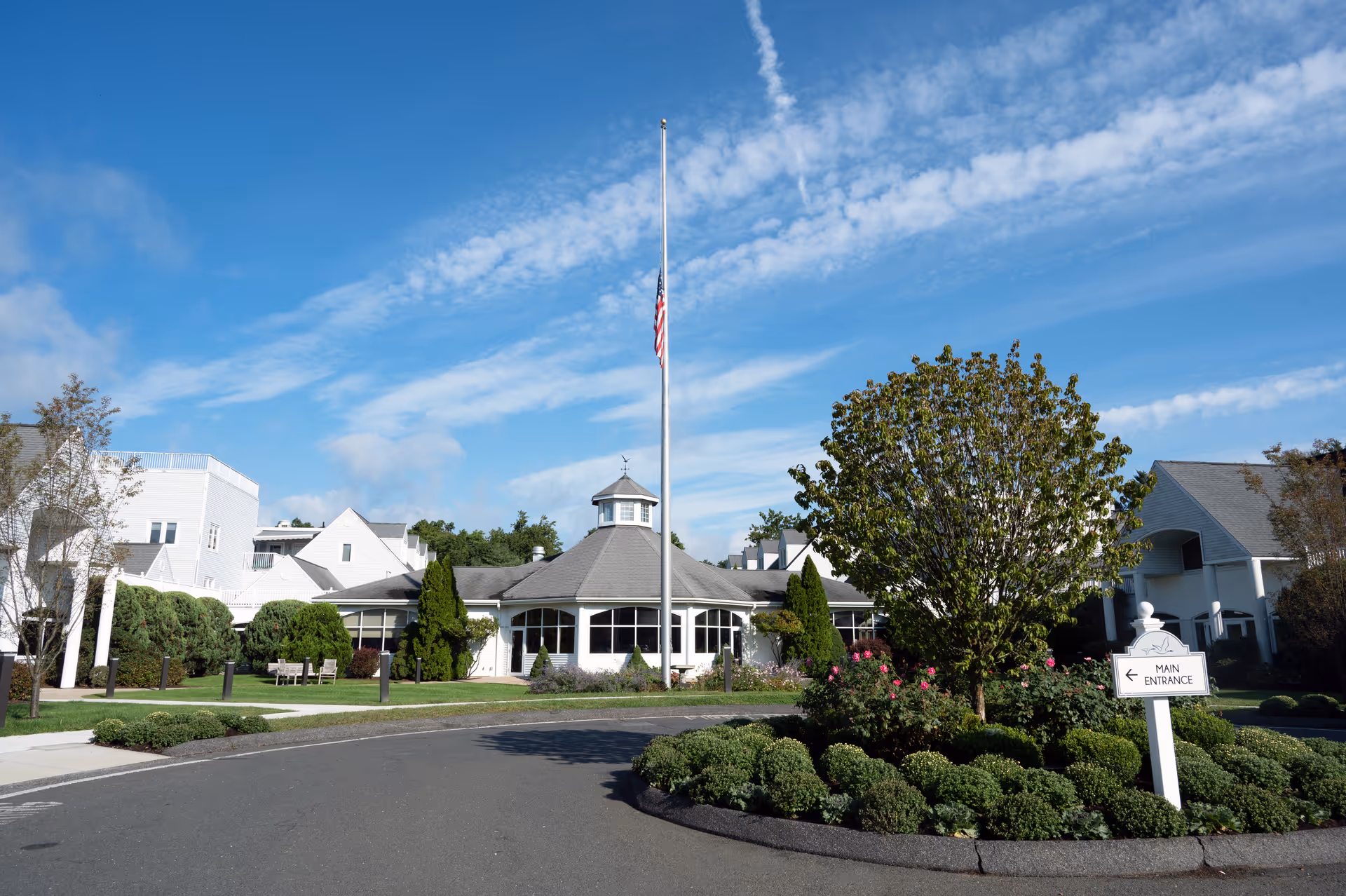 Front exterior of a senior living building with a circular driveway, flagpole at half-mast, and landscaped entrance.