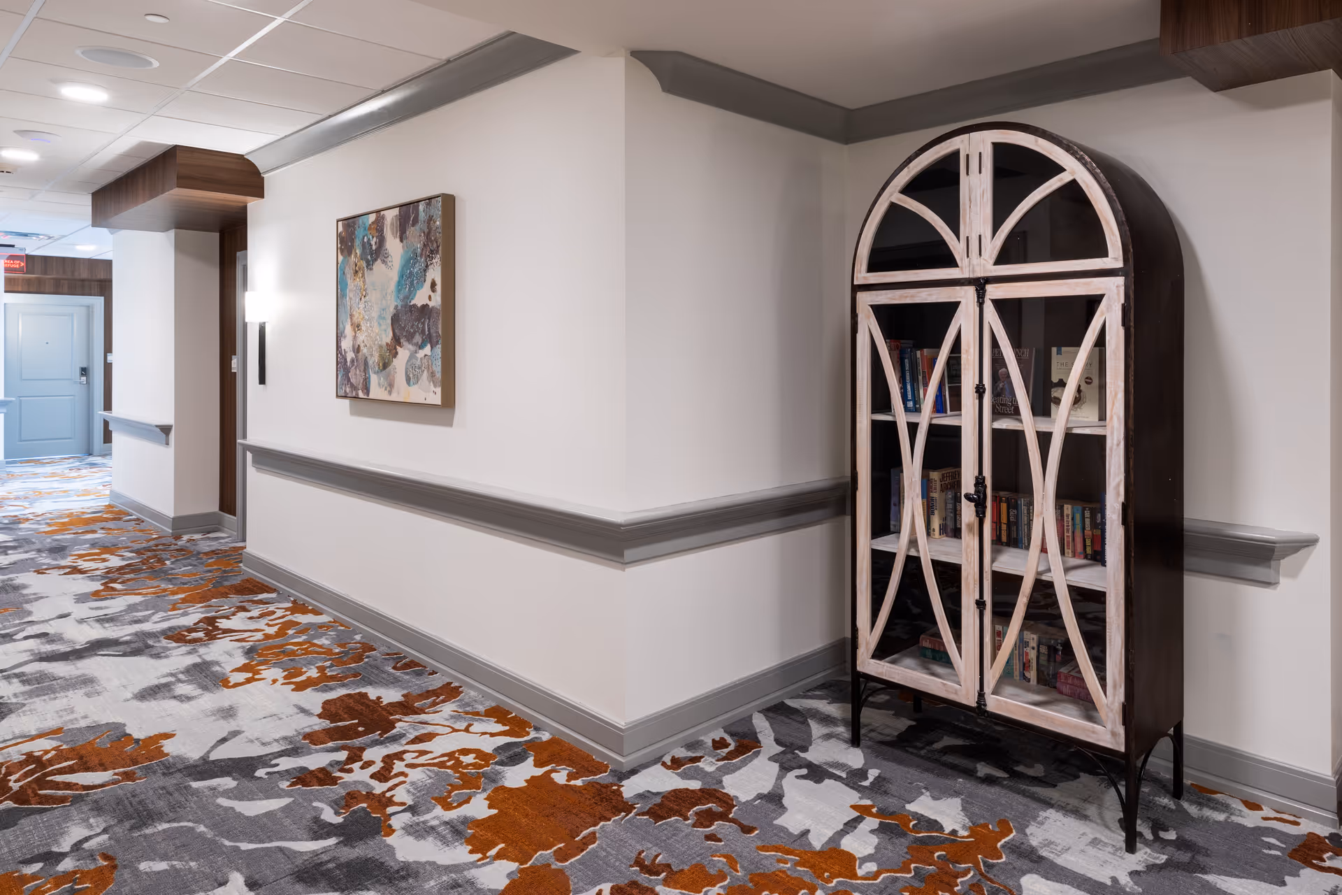 A hallway in a senior living facility with patterned carpet in shades of gray, white, and orange. The walls are painted light gray with a darker gray chair rail. On the right side, there is a decorative wooden cabinet with glass doors containing books. A colorful abstract painting is hung on the wall, and a closed door is visible at the end of the hallway.