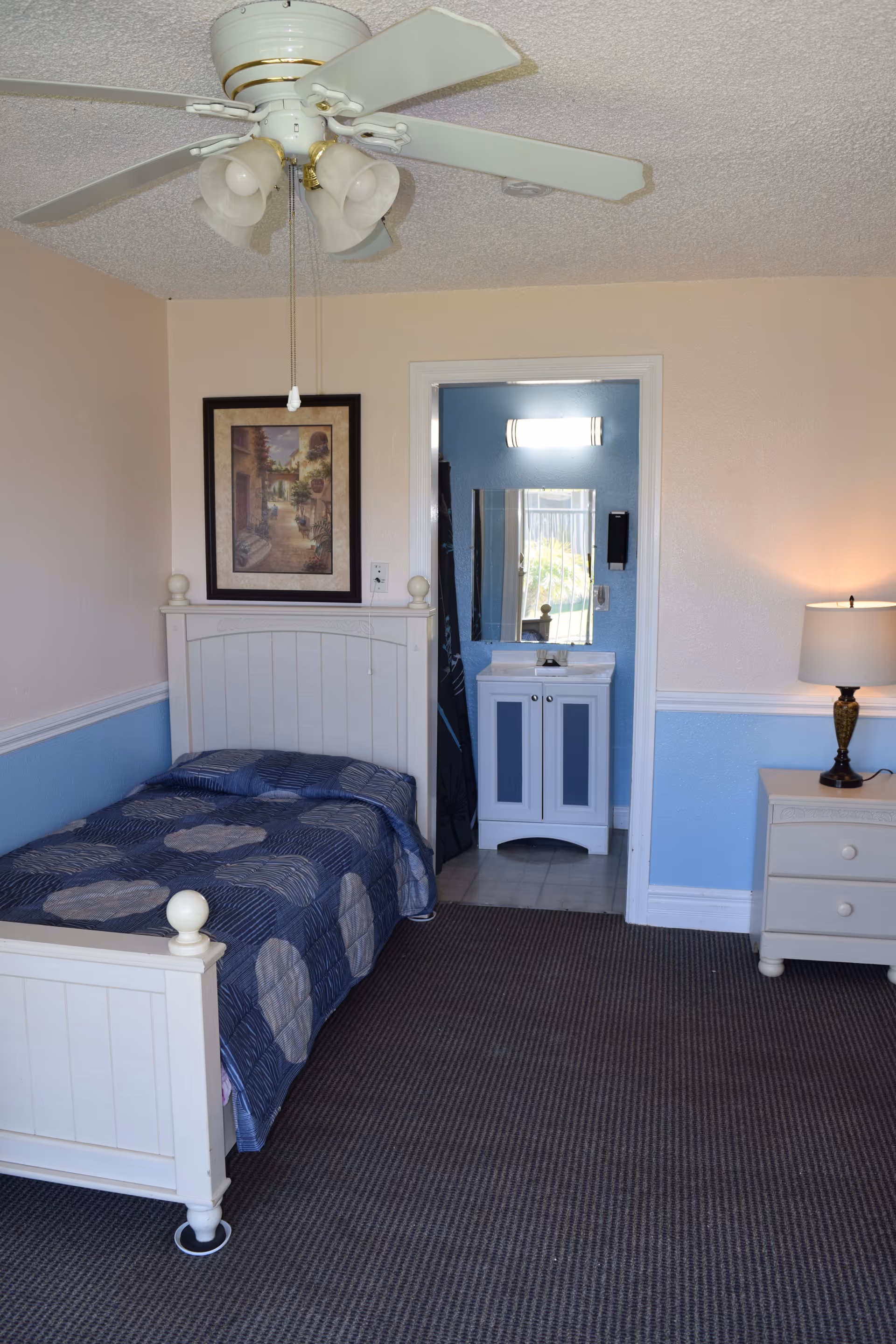 A small bedroom with a white wooden bed frame and a blue patterned bedspread. There is a ceiling fan with lights above the bed. On the wall above the bed is a framed picture. To the right of the bed is a white nightstand with a lamp. An open doorway leads to a small bathroom area with a sink, mirror, and light fixture.