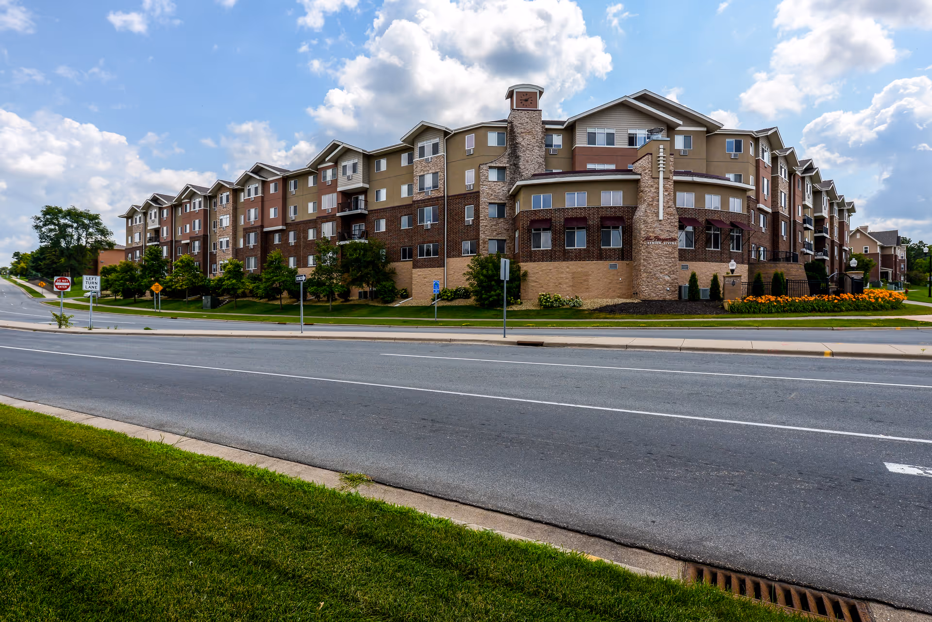Exterior view of a large multi-story senior living facility building with a mix of brick and siding, surrounded by well-maintained landscaping and a road in the foreground under a partly cloudy sky.