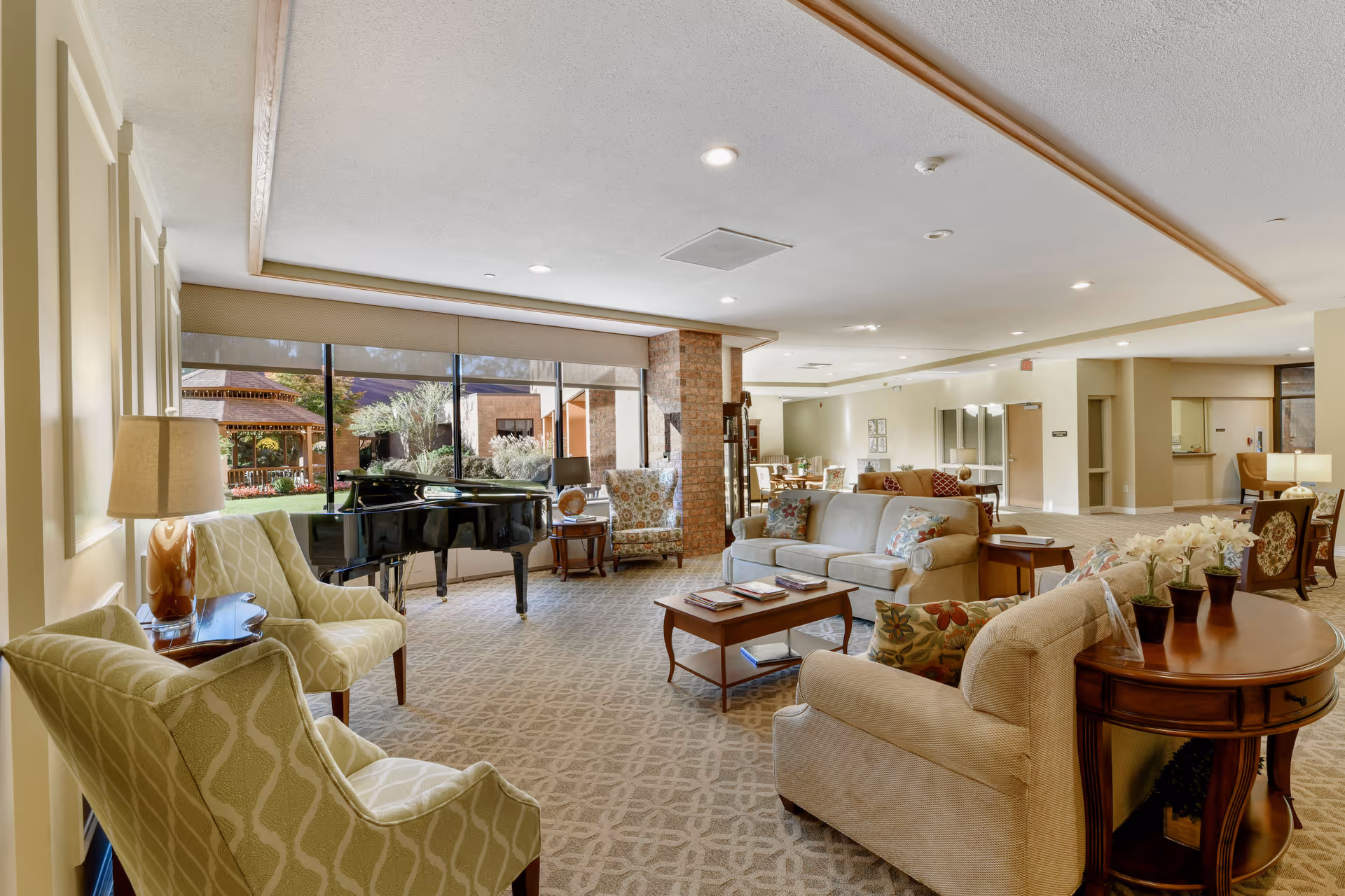 A spacious and well-lit living room area in Worthington Christian Village featuring comfortable upholstered chairs and sofas arranged around wooden coffee tables. A black grand piano is positioned near large windows that offer a view of a garden gazebo outside. The room has beige walls, patterned carpet, and ceiling lights, creating a warm and inviting atmosphere.