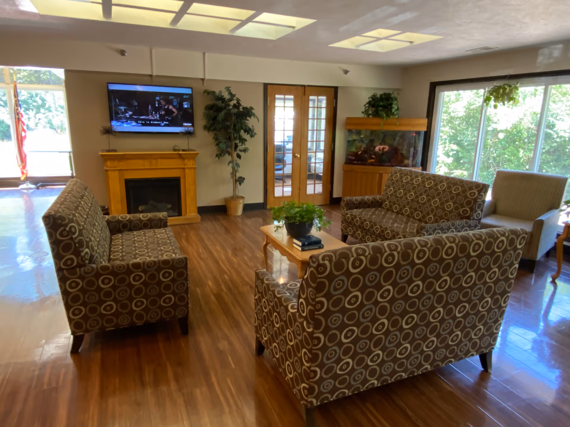 A bright common living room with patterned sofas arranged around a coffee table, a wall-mounted TV, fireplace, fish tank, and large windows.