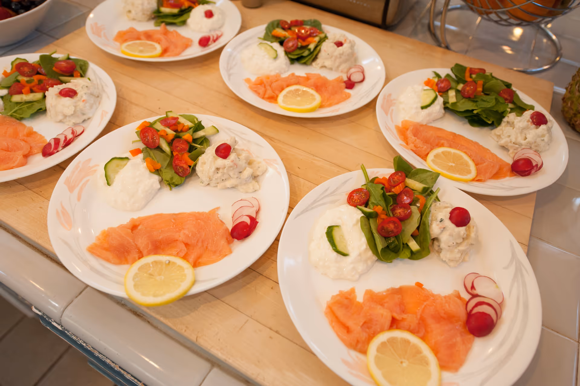 Several plates arranged on a wooden surface, each containing slices of smoked salmon with a lemon wedge, a small salad with cherry tomatoes and cucumber, a dollop of white creamy sauce, and some radish slices.