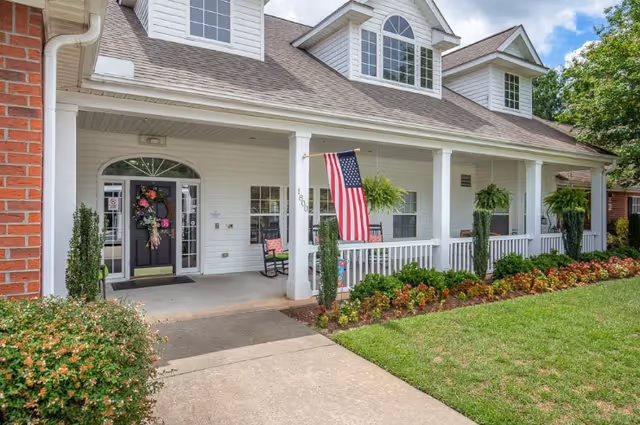 Front exterior view of a white senior living facility building with a covered porch, American flag, hanging plants, and a well-maintained garden with shrubs and flowers. The entrance door is decorated with a floral wreath.