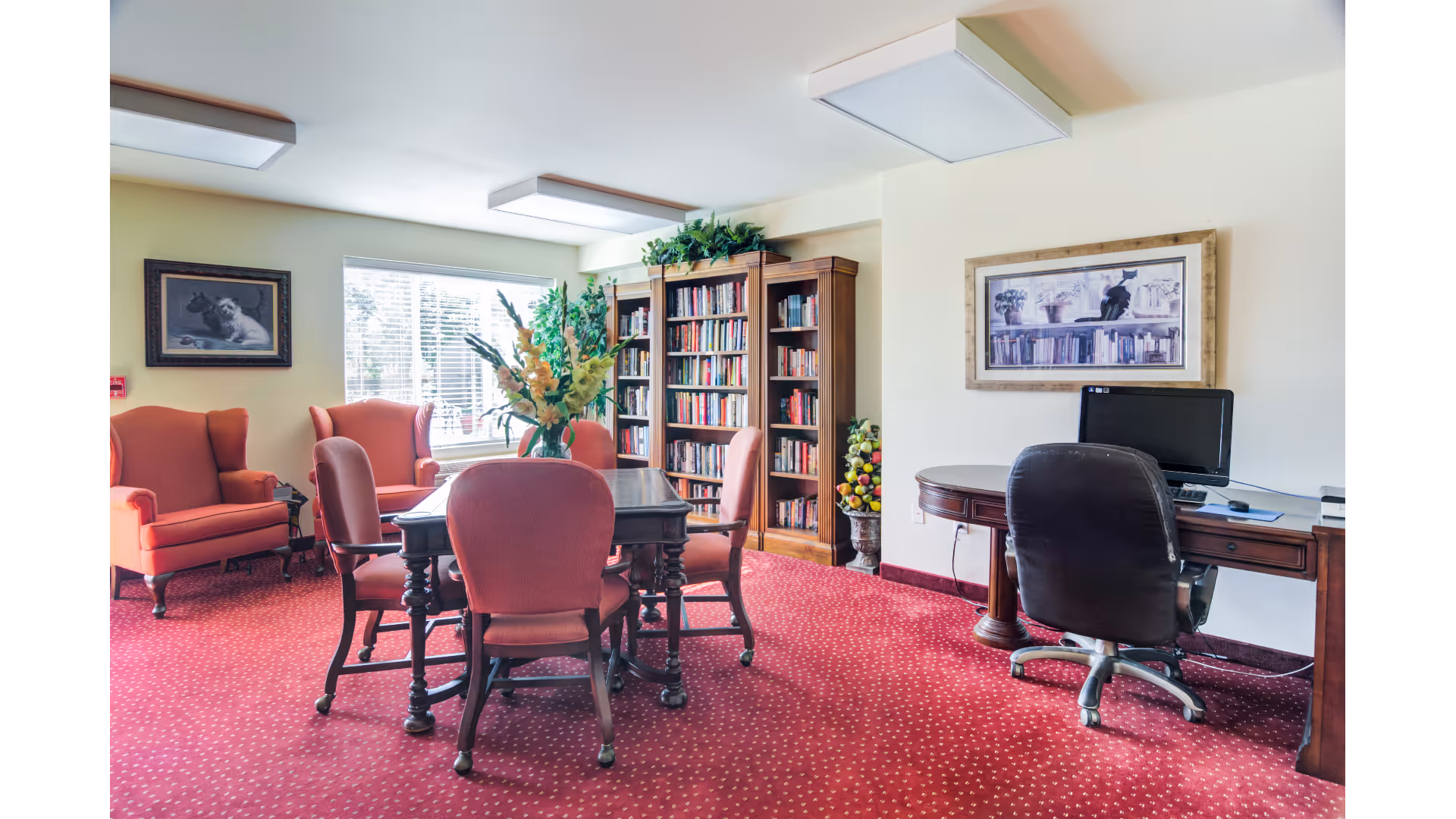 A cozy room with red carpet and matching upholstered chairs around a wooden table. There are two orange armchairs near a window with blinds, a wooden bookshelf filled with books, and a desk with a computer and office chair. The walls have framed artwork, and there are plants on top of the bookshelf and a decorative fruit arrangement beside it.