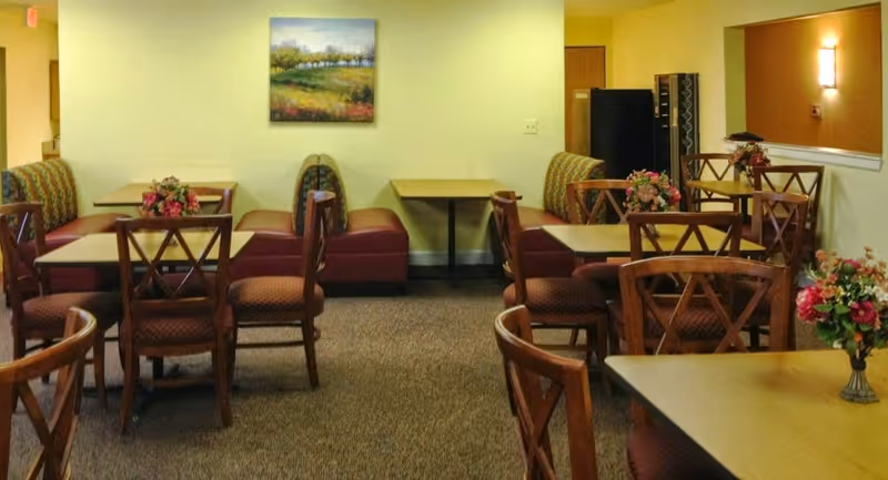 Small dining/common room with multiple tables, wooden chairs, booth seating and floral centerpieces.