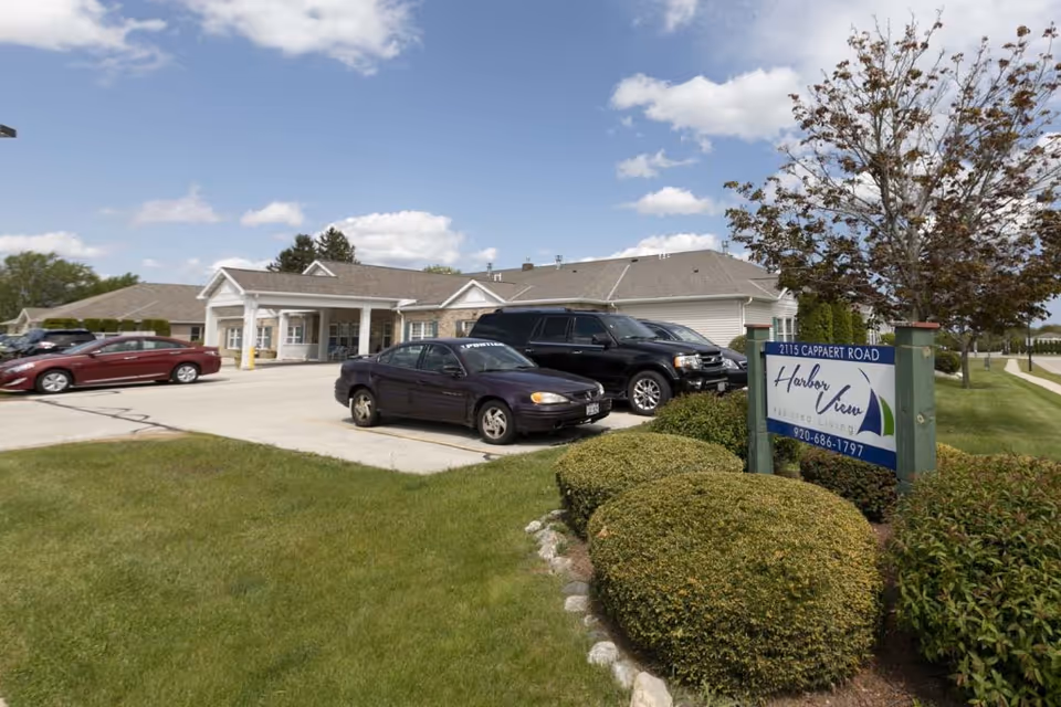 Exterior view of Harbor View Assisted Living facility showing the building entrance, parked cars, manicured bushes, a tree, and a sign with the facility name and contact information under a partly cloudy sky.