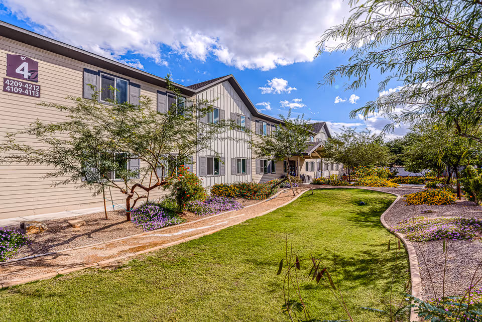 Exterior view of a two-story senior living facility building with beige siding and multiple windows. The building is surrounded by a well-maintained garden with green grass, flowering plants, and small trees under a partly cloudy blue sky.