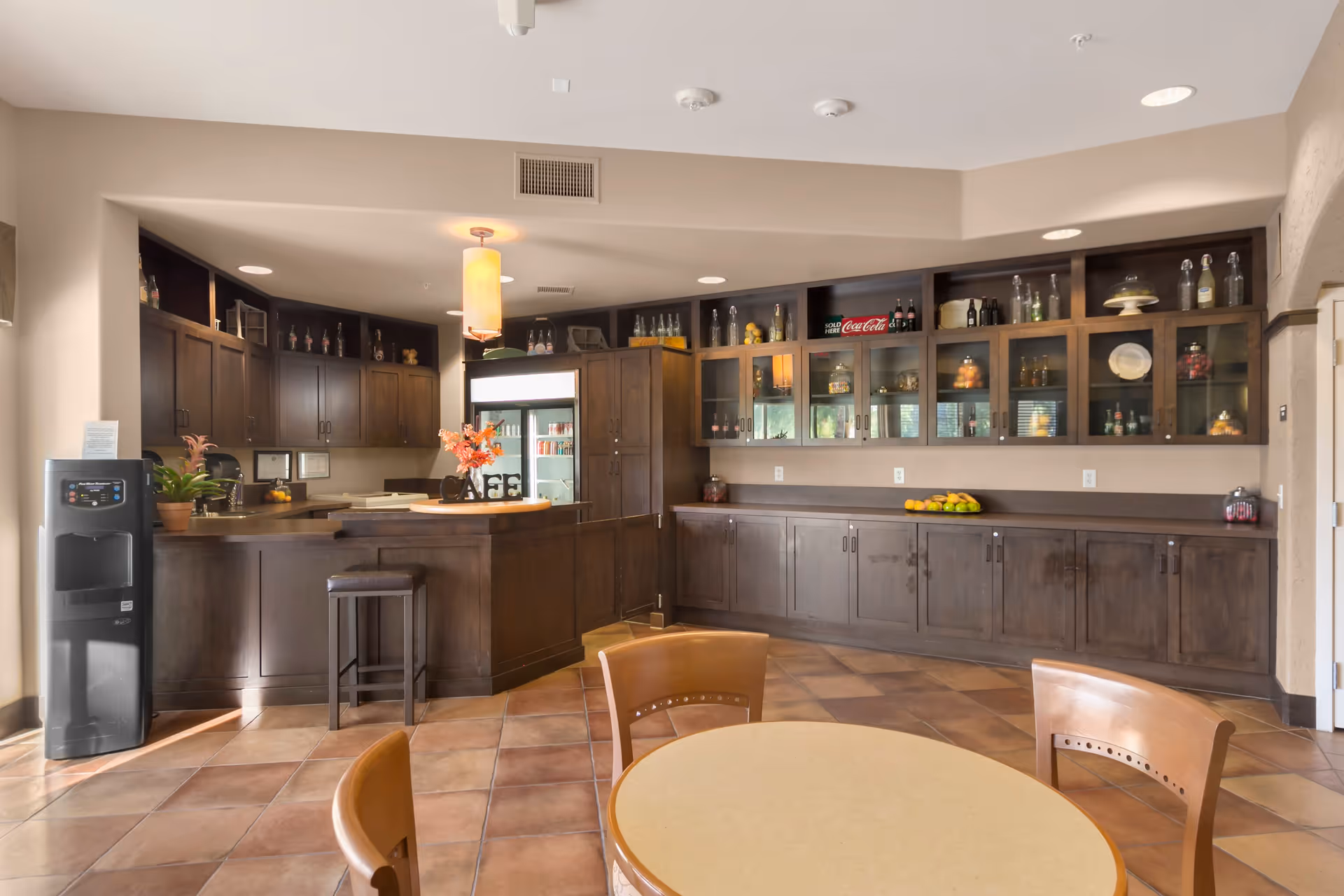 Interior view of a senior living facility's kitchen and dining area with dark wood cabinets, a counter with a stool, a round table with chairs, a water dispenser, and decorative items on shelves and countertops.