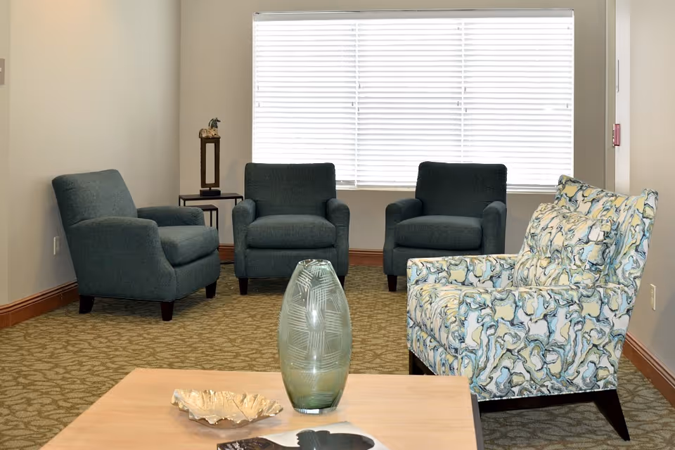 A seating area with four upholstered armchairs around a coffee table holding a decorative vase in front of a large window with blinds.