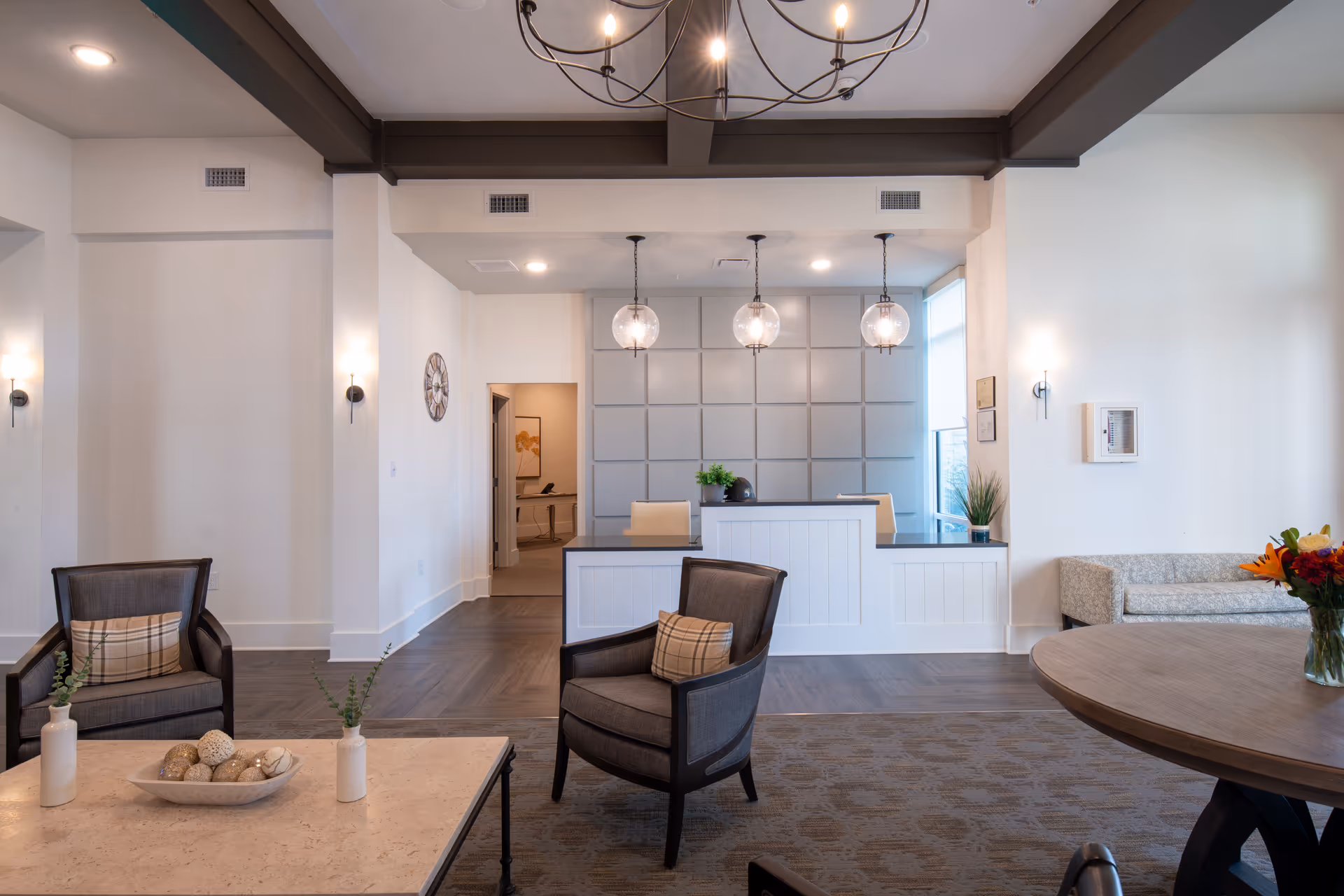 A modern and well-lit reception area with a white front desk, three hanging globe pendant lights, and a decorative wall panel behind the desk. The room features comfortable seating including two dark armchairs with plaid pillows, a round wooden table with a vase of flowers, and a light-colored bench along the wall. The floor has a patterned carpet and wood flooring, and the ceiling has exposed dark beams with a chandelier.