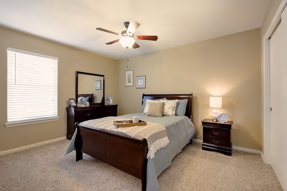 A cozy bedroom with a wooden bed frame, blue bedding, and decorative pillows. There is a wooden dresser with a mirror and framed pictures on the left side, and a wooden nightstand with a lamp and a decorative plate on the right side. The room has beige walls, a ceiling fan with lights, a window with blinds, and carpeted flooring.
