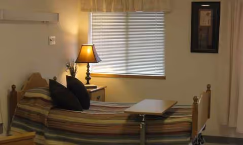 Bed with striped bedding, nightstand, lamp, and window blinds in a bedroom.