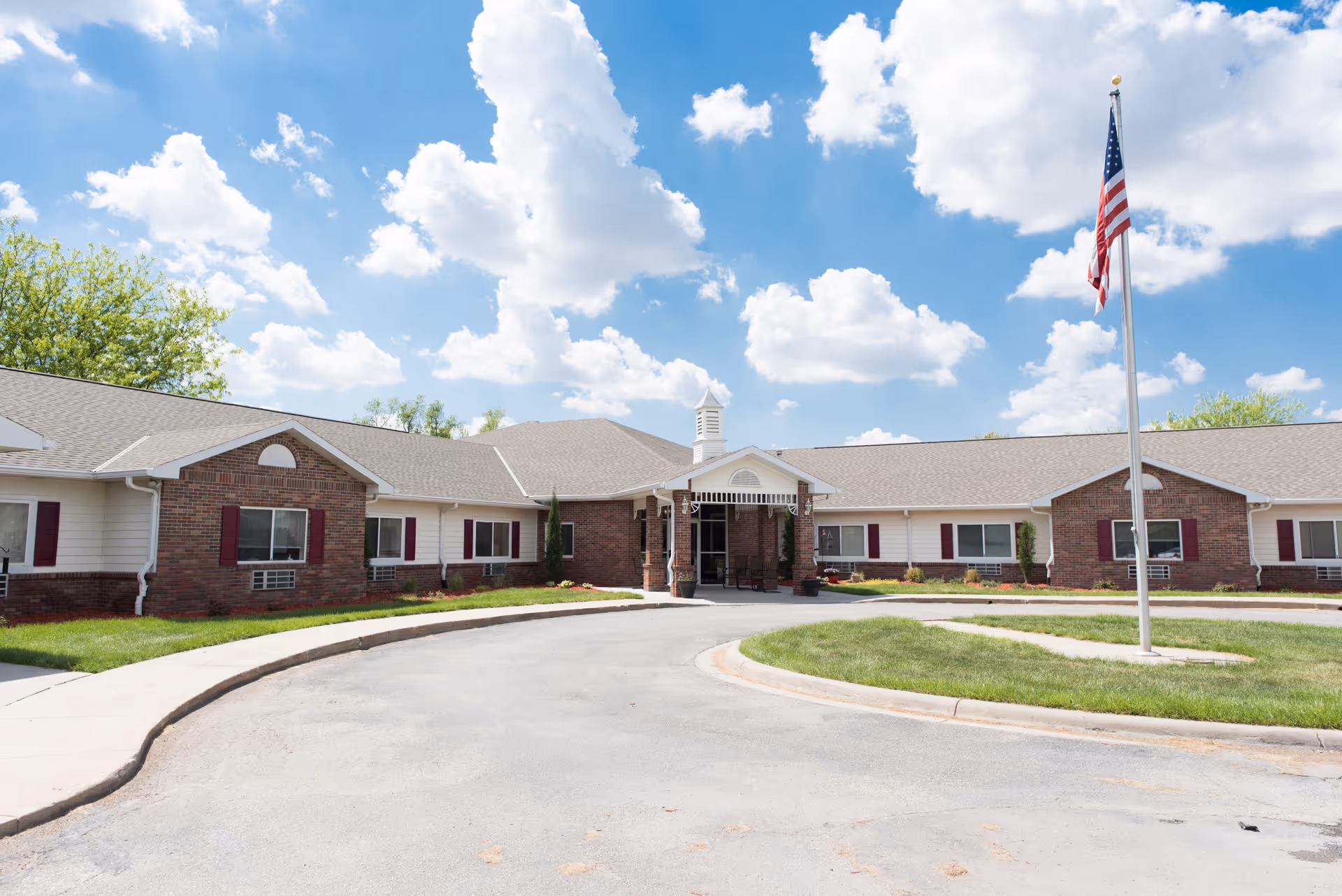 Exterior view of a single-story assisted living facility building with brick and white siding, a covered entrance, and an American flag on a flagpole in front. The sky is blue with scattered clouds.