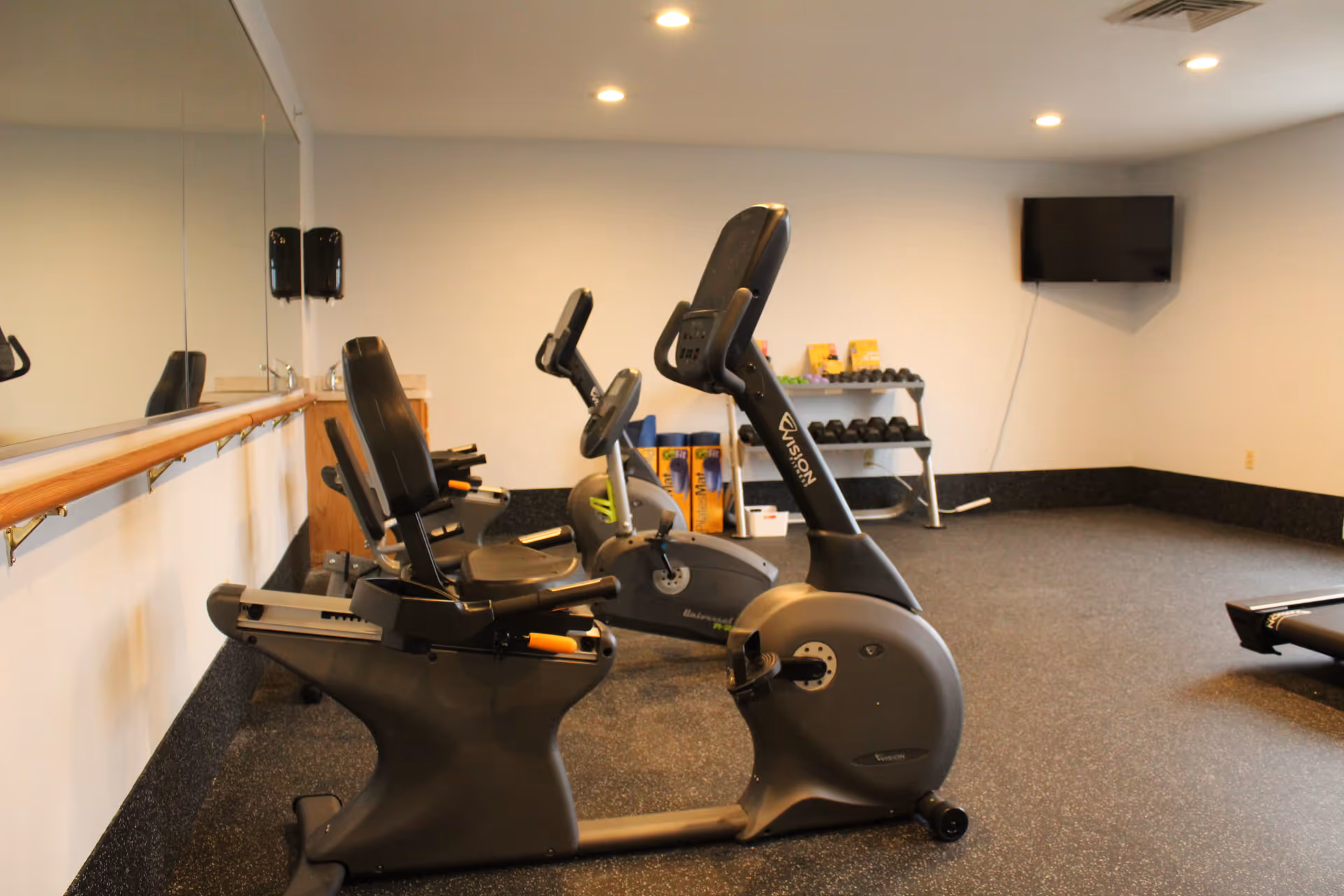 Small fitness room with recumbent exercise bikes, a mirror, dumbbells and a wall-mounted TV.