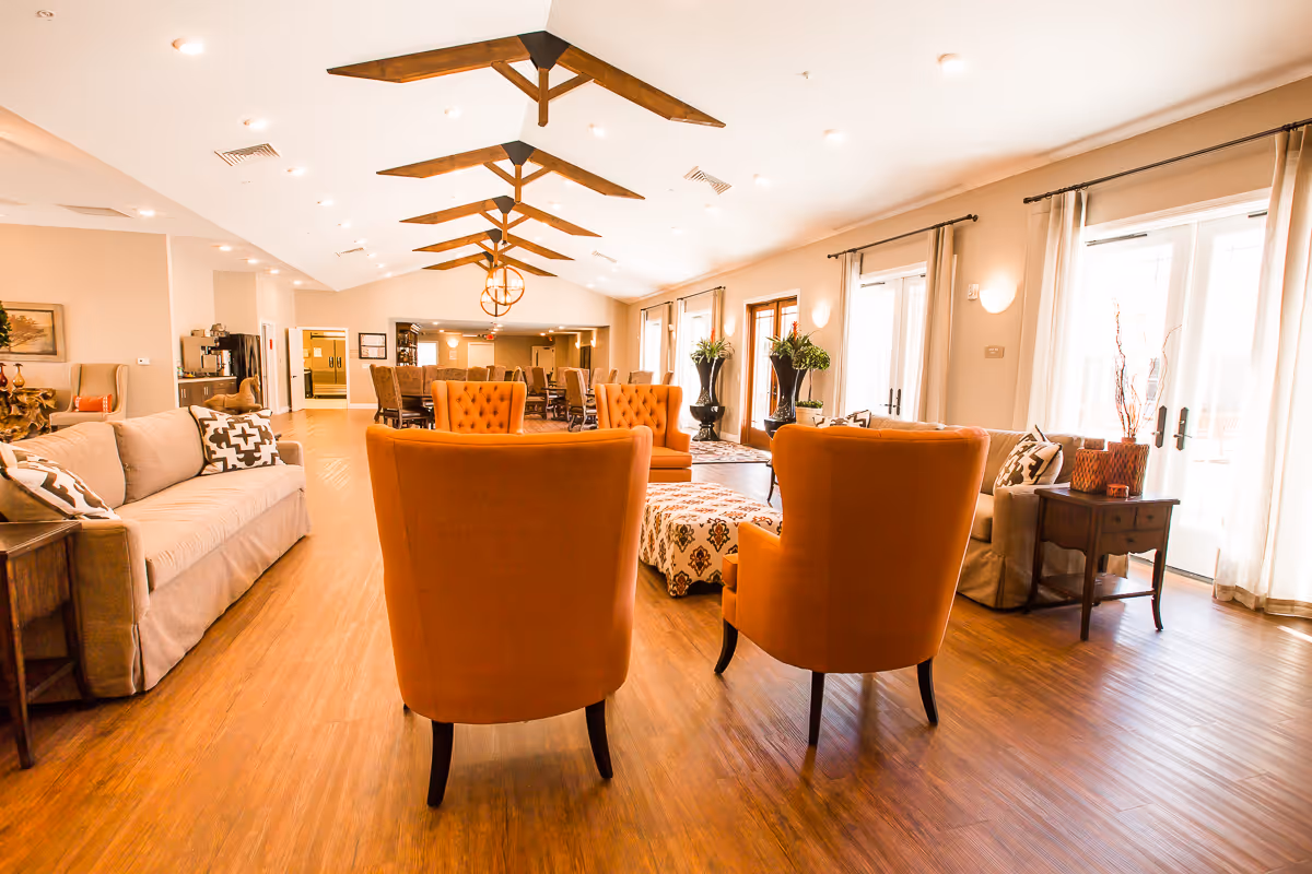 Spacious and well-lit senior living common area with wooden flooring, orange armchairs, beige sofas, patterned ottoman, large windows with curtains, and wooden ceiling beams with modern light fixtures.