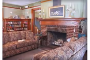 A cozy living room with two patterned sofas facing each other, a brick fireplace with a decorative mantel, a framed floral painting above the fireplace, and a bookshelf filled with books in the background.