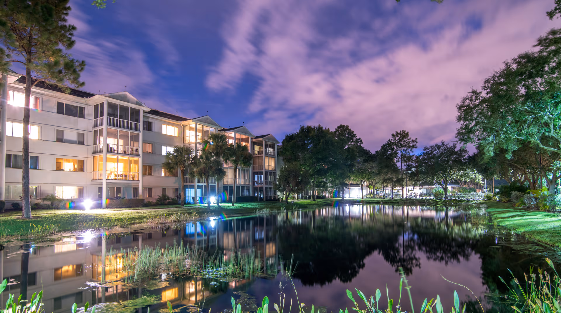 Nighttime view of a multi-story senior living facility building with illuminated windows, reflected in a calm pond surrounded by trees and greenery under a partly cloudy sky.