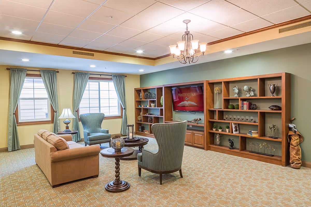 A cozy senior living facility common area with a beige sofa, two green upholstered armchairs, a round wooden coffee table, and a side table with a lamp. The room has large windows with green curtains, a green accent wall with wooden built-in shelves displaying decorative items, and a flat-screen TV mounted in the center. The ceiling features recessed lighting and a chandelier.