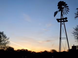 Silhouette of a tall windmill structure against a sunset sky with trees in the foreground.