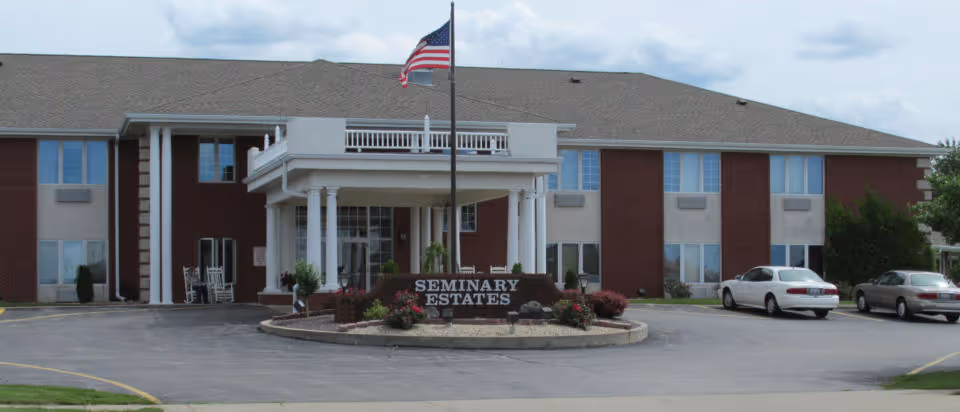 Front entrance of a two-story brick senior living building with a flagpole and a sign reading 'Seminary Estates'.