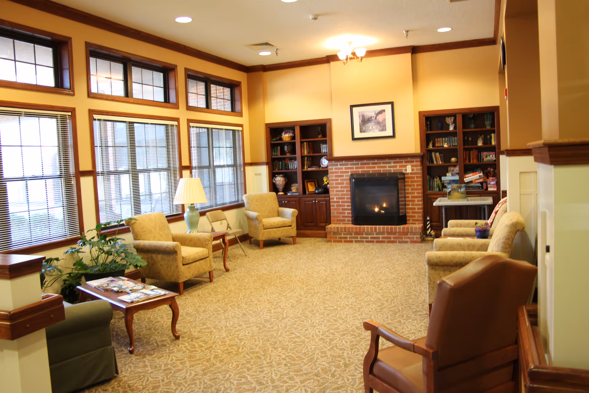 Cozy common room with armchairs, a brick fireplace flanked by bookshelves, and large windows along one wall.