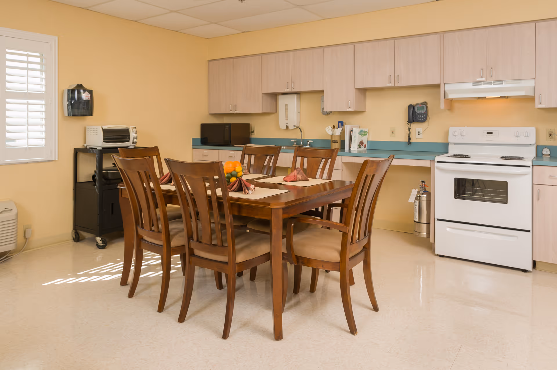A kitchen dining area with a wooden table and six chairs. The table is set with placemats, napkins, and a bowl of fruit. The kitchen has light wood cabinets, a white stove, a microwave, a toaster oven, and a wall-mounted phone. The walls are painted yellow and there is a window with white shutters letting in natural light.