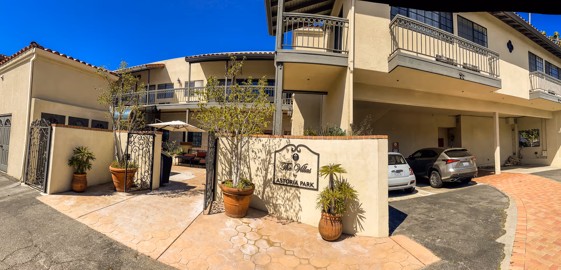 Exterior view of The Villas by Astoria Park facility showing a gated entrance with potted plants, a seating area with an umbrella inside the gate, and a parking area with cars on the right side under a covered section of the building.