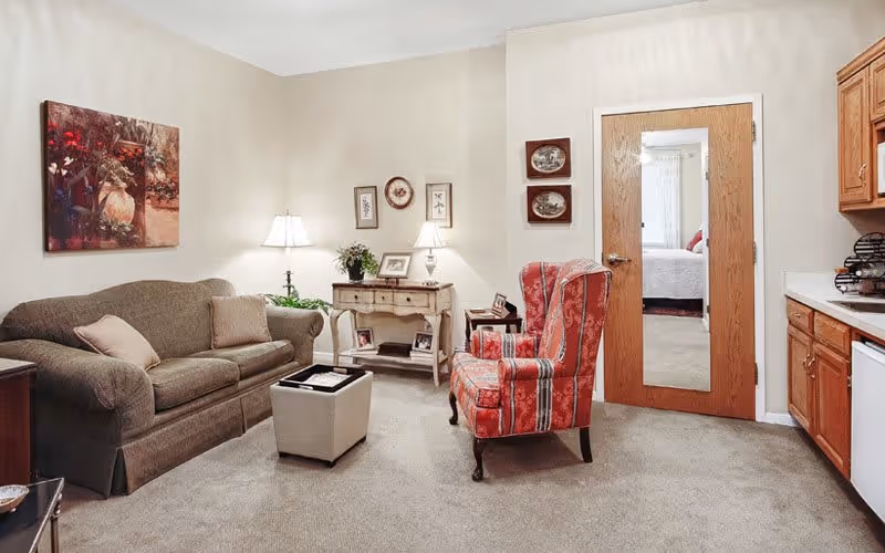A cozy living room featuring a brown sofa with two cushions, a red patterned armchair, a small ottoman, and a wooden side table with two lamps and decorative items. There is a door with a full-length mirror leading to a bedroom, and a partial view of a kitchen area with wooden cabinets and a dishwasher.