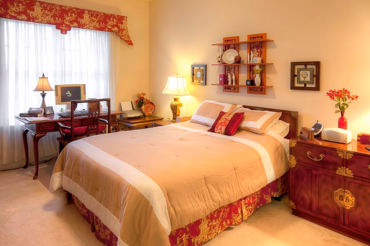 A cozy bedroom with a large bed covered in beige and white bedding with red decorative pillows. To the left, there is a wooden desk with a computer, lamp, books, and a chair. On the right side of the bed, there is a wooden nightstand with a lamp, a red vase with flowers, a clock, and a small radio. The walls are decorated with framed pictures and a wooden shelf holding decorative items. A window with sheer white curtains and a red valance is behind the desk.