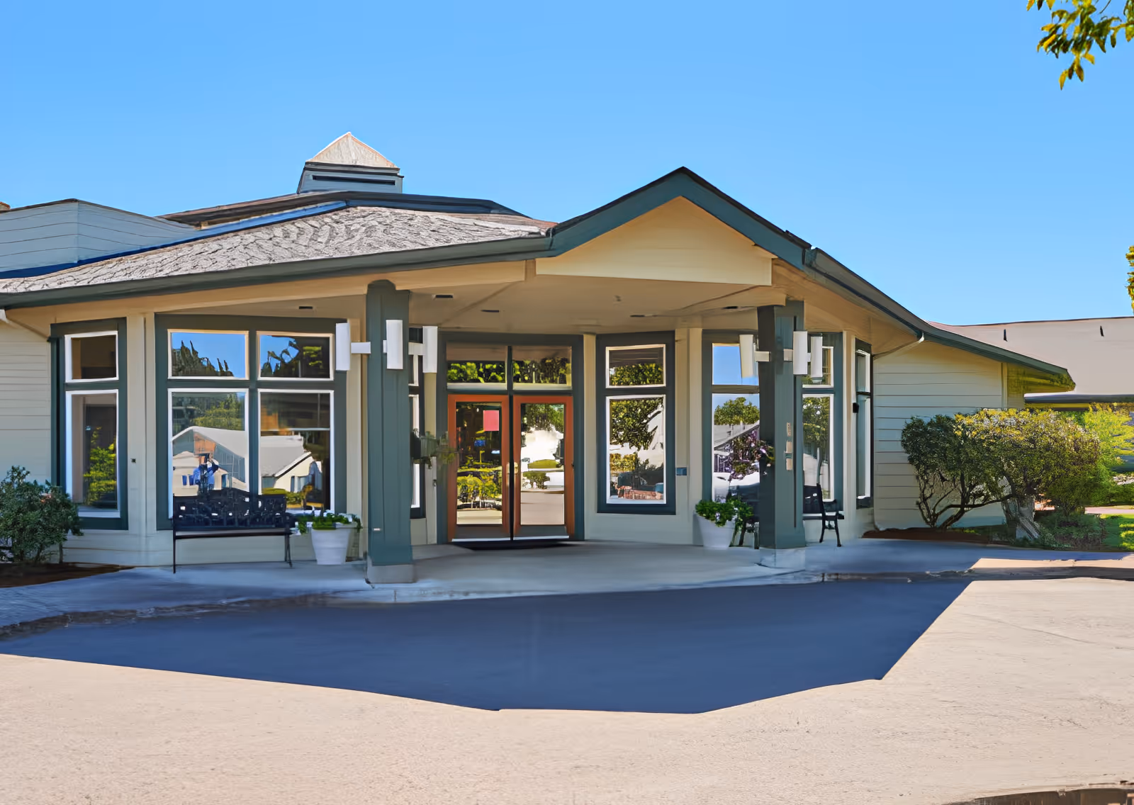 Entrance of Elliott Residence featuring a covered driveway with a peaked roof, large windows, double glass doors, outdoor benches, potted plants, and surrounding greenery under a clear blue sky.