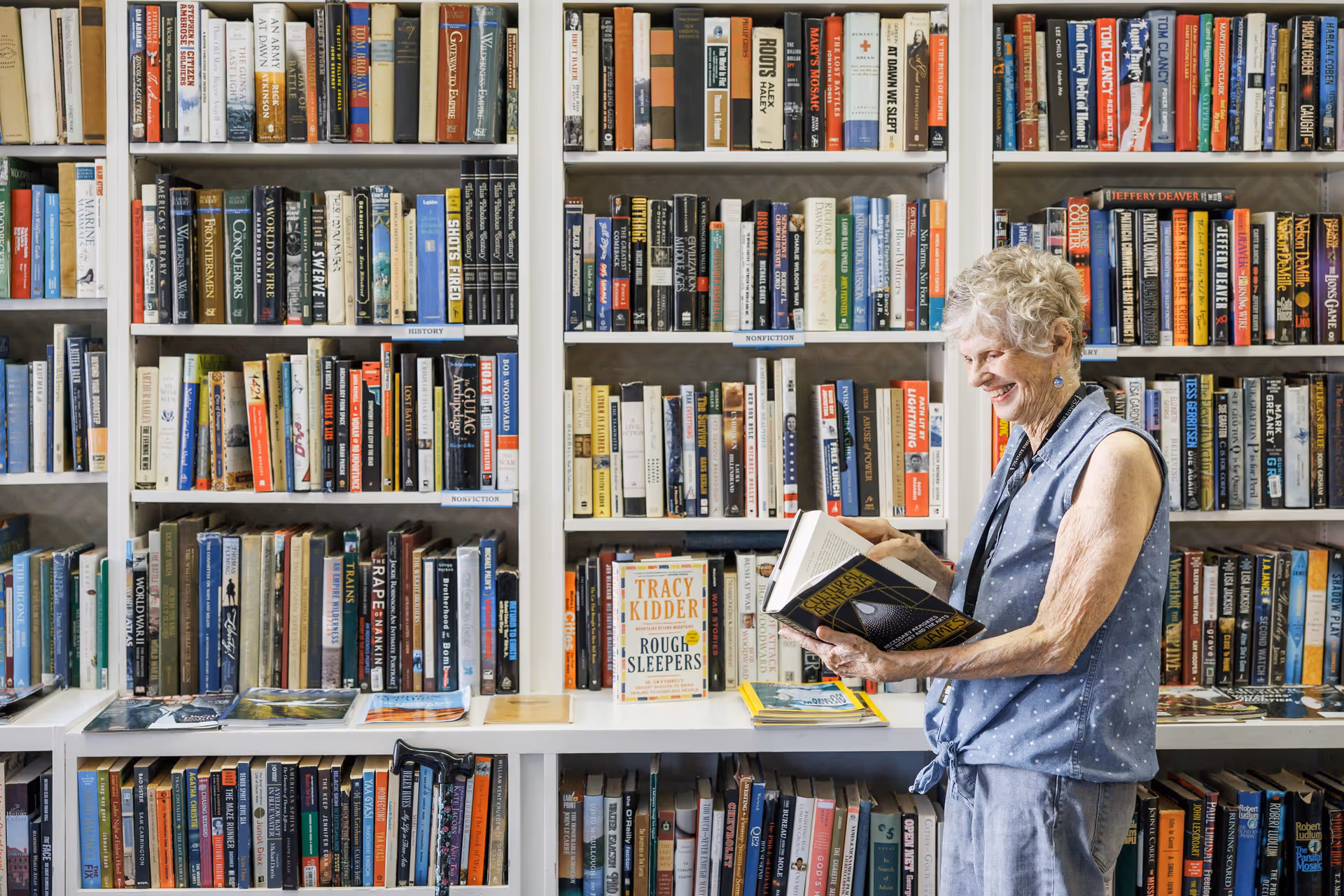 An elderly woman with short gray hair wearing a sleeveless blue blouse and light pants is standing in front of a large bookshelf filled with various books. She is smiling and reading a book while holding it open in her hands. The bookshelf is organized with books of different sizes and colors, and some books are displayed facing forward on a white shelf.