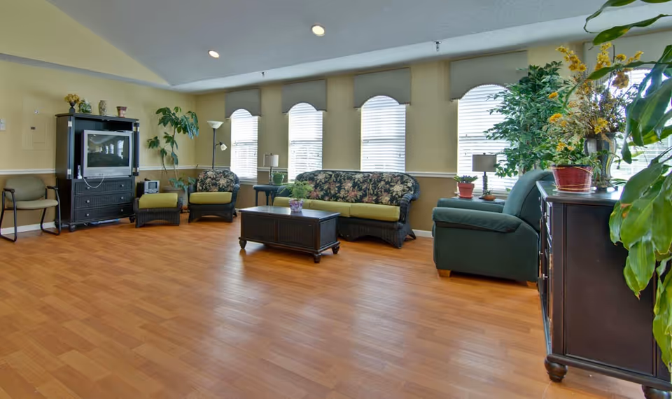 A spacious living room with wooden flooring, featuring a floral-patterned sofa, two matching armchairs, a green armchair, a wooden coffee table, and a TV cabinet with a television. The room has several large windows with blinds, potted plants, and soft lighting from lamps.