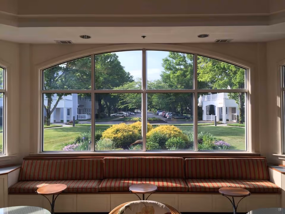 Interior view of a seating area with a long cushioned bench with red and beige striped upholstery beneath a large arched window. Outside the window, there is a landscaped garden with bushes, trees, and a parking area with cars. The room has soft natural lighting.