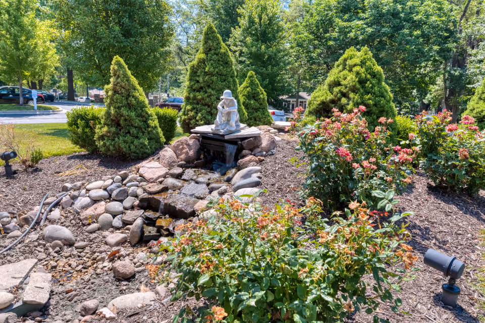 A landscaped garden area with a small water feature surrounded by rocks and mulch. There is a statue of a person sitting on a platform above the water. The garden includes green shrubs, flowering plants with pink blooms, and several conical evergreen trees. In the background, there are parked cars and more trees under a clear blue sky.