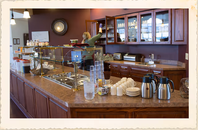 A kitchen or serving area with a countertop displaying cups, plates, pitchers, and coffee thermoses. A woman is reaching into a glass-front cabinet mounted on a purple wall. The countertop has a glass sneeze guard and various kitchen appliances are visible in the background.