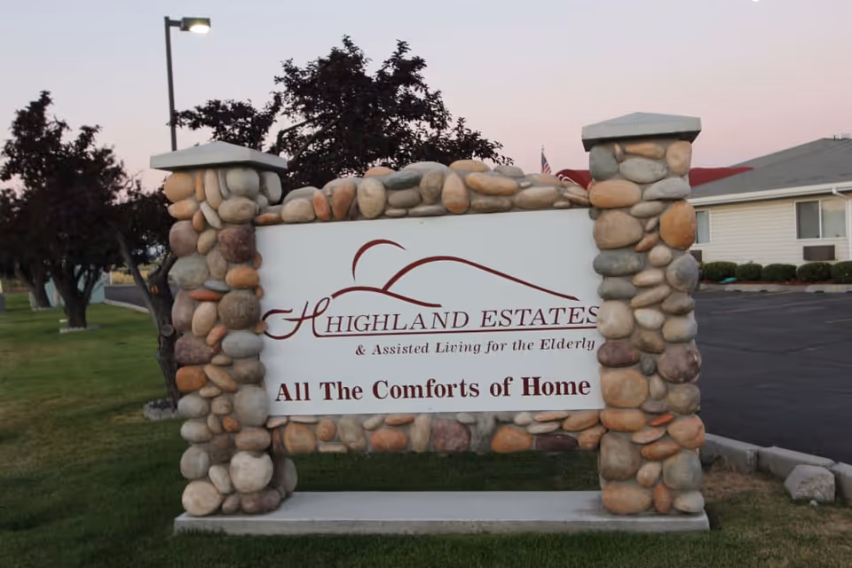 Stone-framed sign for Highland Estates Assisted Living & Memory Care, reading 'Highland Estates & Assisted Living for the Elderly All The Comforts of Home', set on a grassy area with trees and a building in the background during dusk.