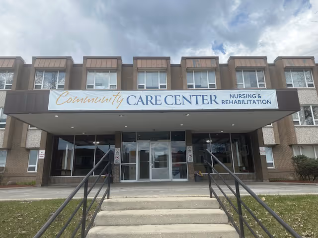Front exterior view of a three-story brick building with a large sign above the entrance that reads 'Community Care Center Nursing & Rehabilitation'. There are steps and handrails leading up to glass double doors under a covered entryway.