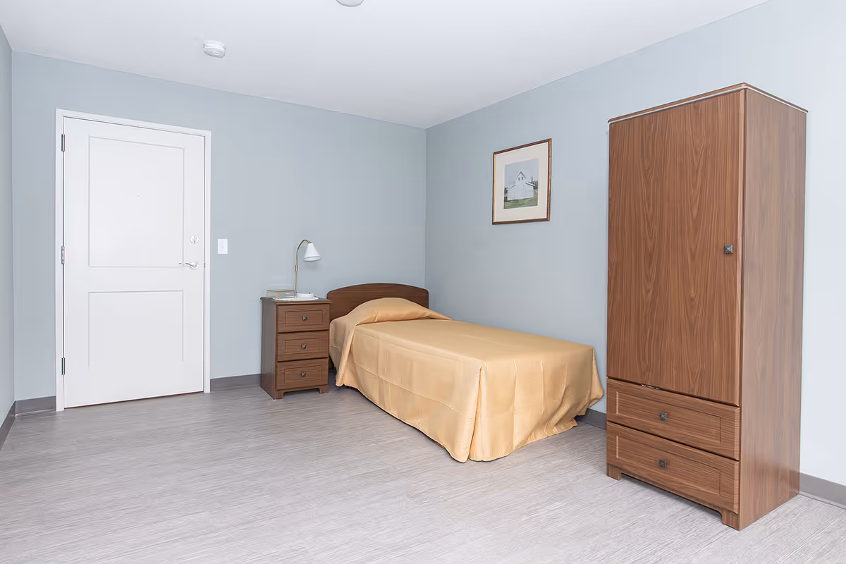 Small bedroom with a single bed covered in a beige bedspread, a wooden nightstand with a lamp, and a tall wooden wardrobe against pale blue walls.