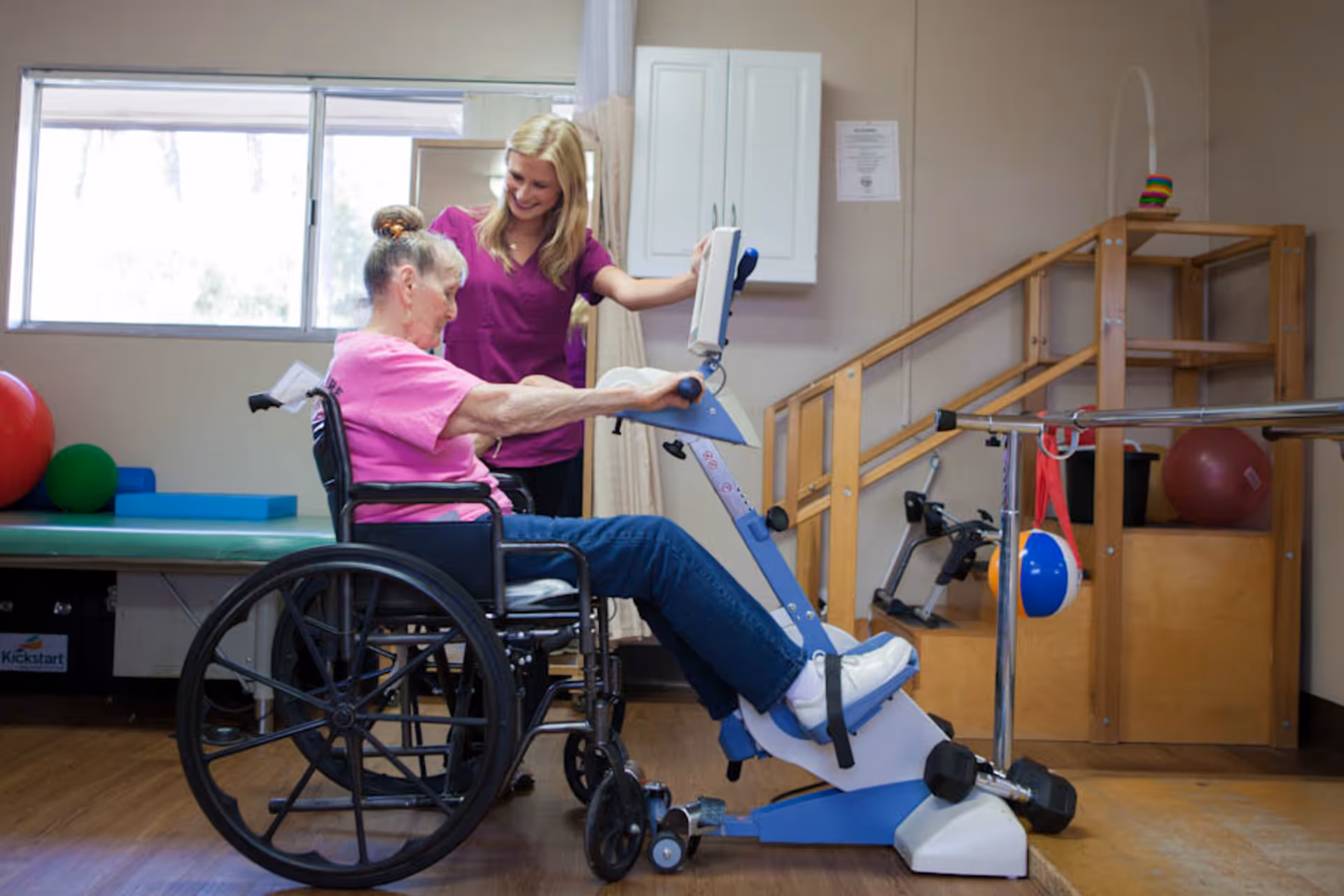 An elderly woman in a wheelchair using a pedal exerciser machine with assistance from a smiling female caregiver in a purple uniform in a therapy room equipped with exercise balls and parallel bars.