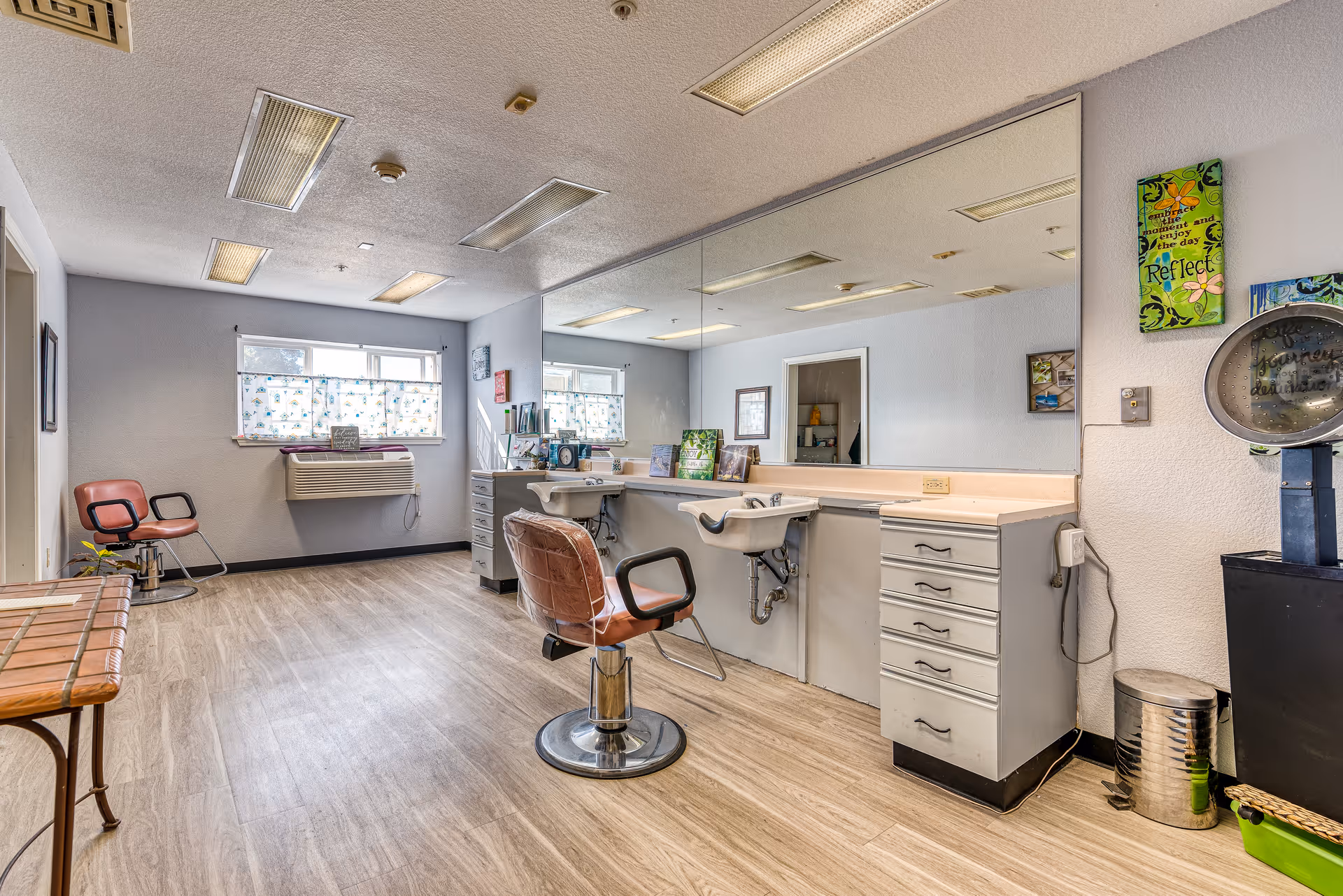 Interior view of a salon area in a senior living facility with a large mirror on the wall, two salon chairs, a sink for washing hair, and various decorations on the walls. The room has wood-style flooring and fluorescent ceiling lights.