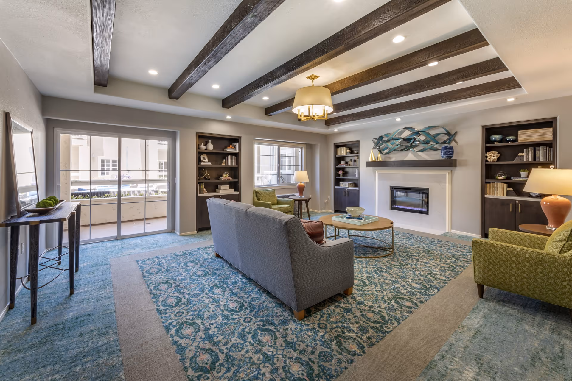 A spacious living room with a blue patterned carpet, gray sofa, green armchairs, a round wooden coffee table, built-in dark wood bookshelves, a modern fireplace with decorative art above it, exposed wooden ceiling beams, and a large sliding glass door leading to an outdoor area.