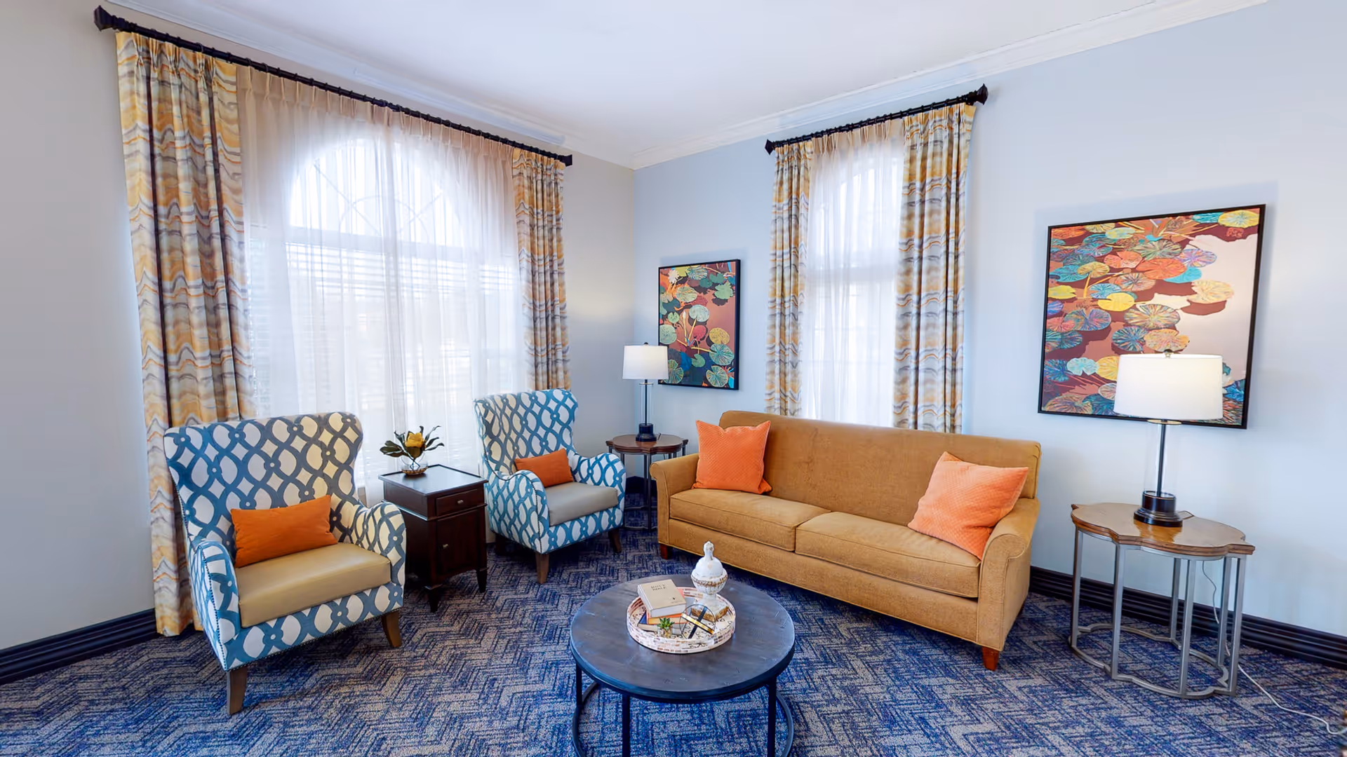 A cozy living room with a mustard yellow sofa adorned with two orange pillows, two blue and white patterned armchairs each with an orange pillow, a round coffee table with decorative items, two side tables with lamps, and two large windows with patterned curtains. The walls are light blue with colorful artwork hanging on them, and the floor is covered with a blue patterned carpet.