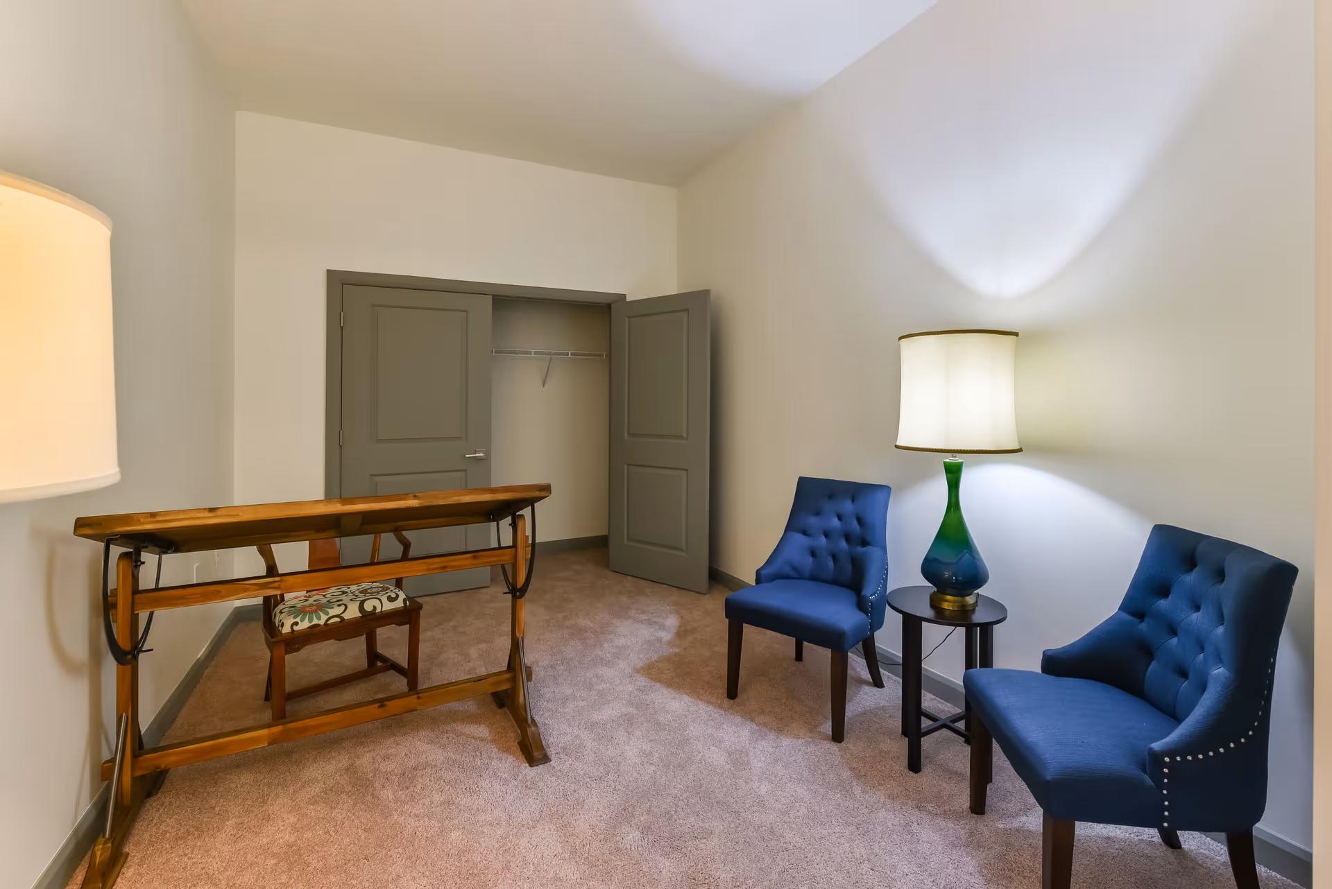 A small room with beige carpet and white walls featuring two blue upholstered chairs with button tufting and nailhead trim, a small round black side table with a green and blue glass lamp, a wooden drafting table with a floral cushioned chair behind it, and an open closet with double gray doors.