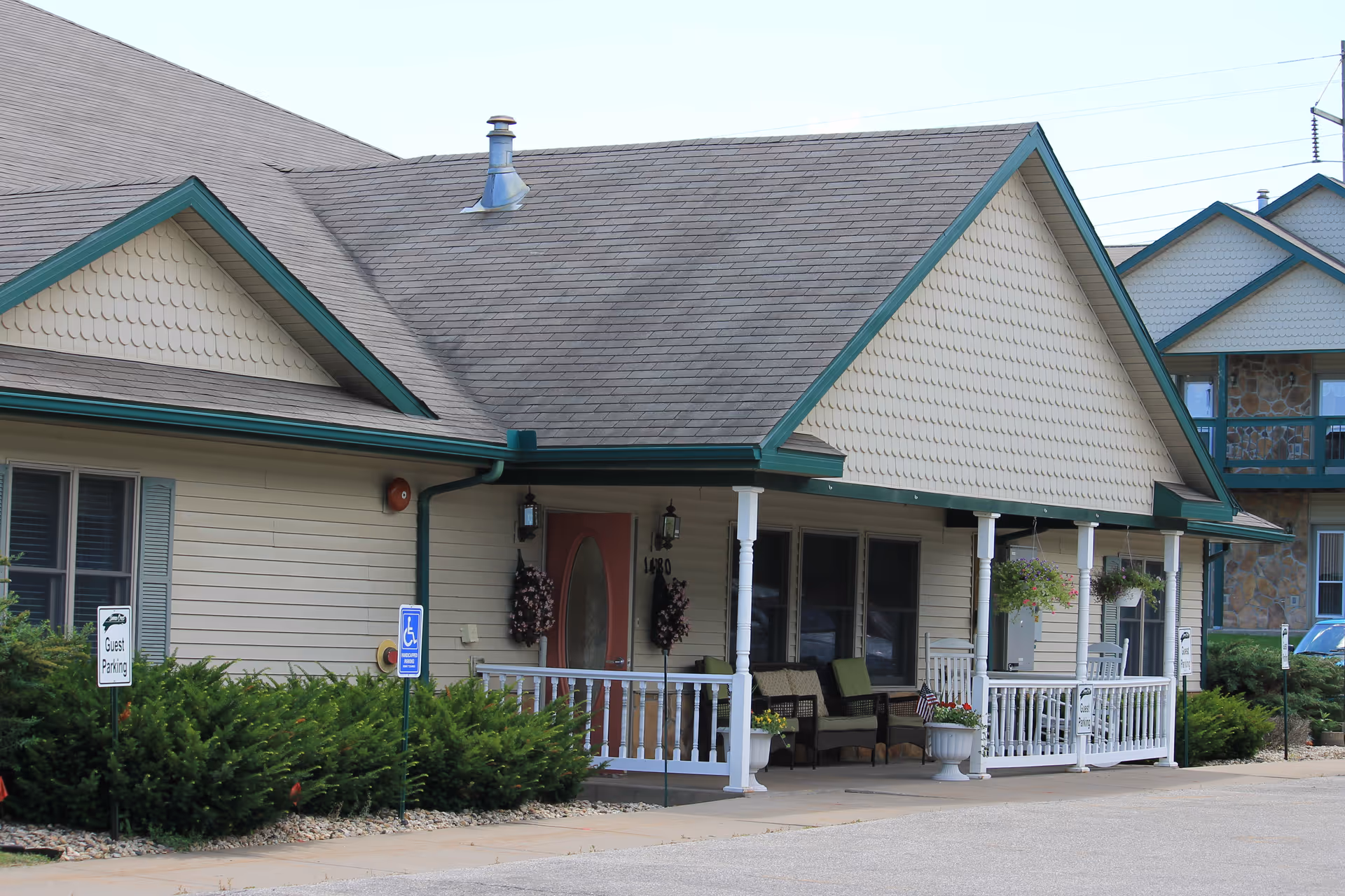 Exterior view of Sienna Crest Assisted Living, Inc. showing a single-story building with a covered porch, white railings, outdoor seating, and hanging flower baskets. There are signs for guest parking and handicapped parking near the entrance.