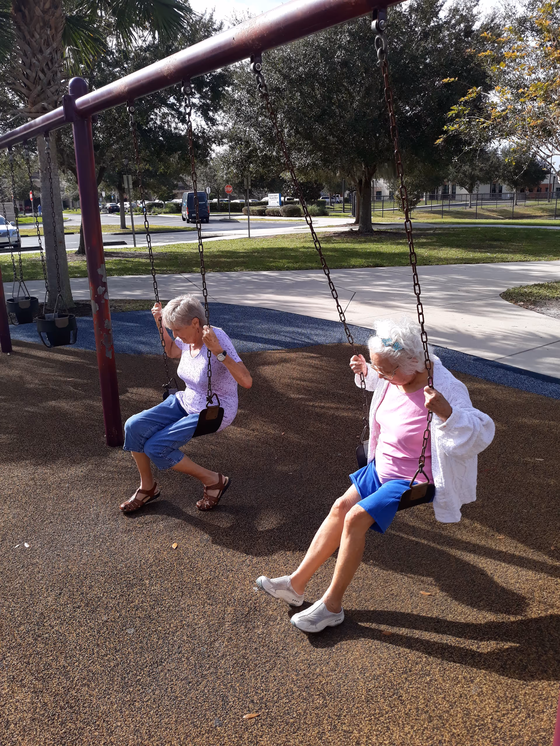 Two older adults sitting on swings at a playground under trees with a parking area in the background.