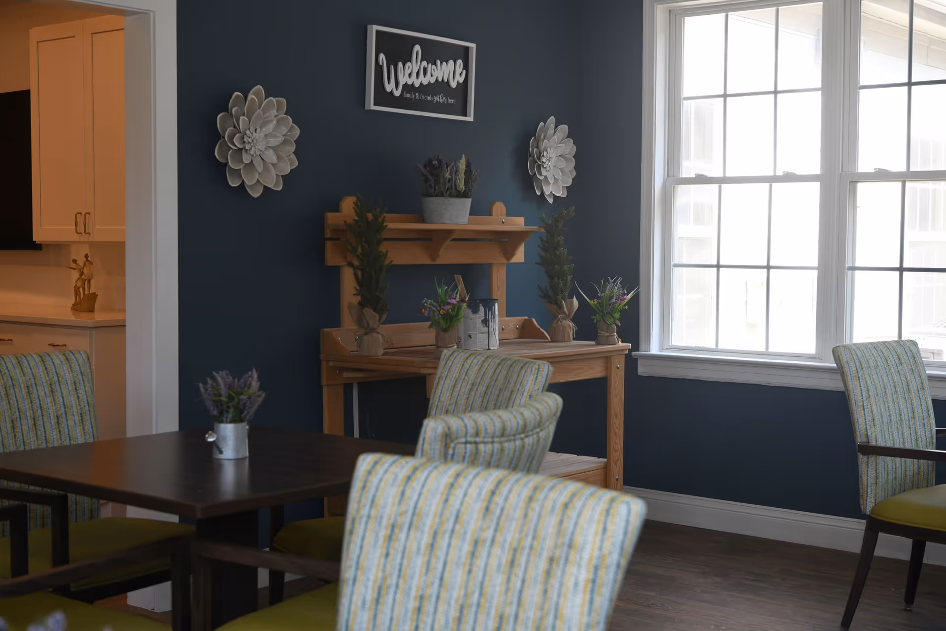 A dining area with a dark wooden table surrounded by striped cushioned chairs. A wooden shelf against a dark blue wall holds small potted plants and decorative items. Above the shelf is a sign that says 'Welcome' and two flower-shaped wall decorations. Large windows let in natural light.