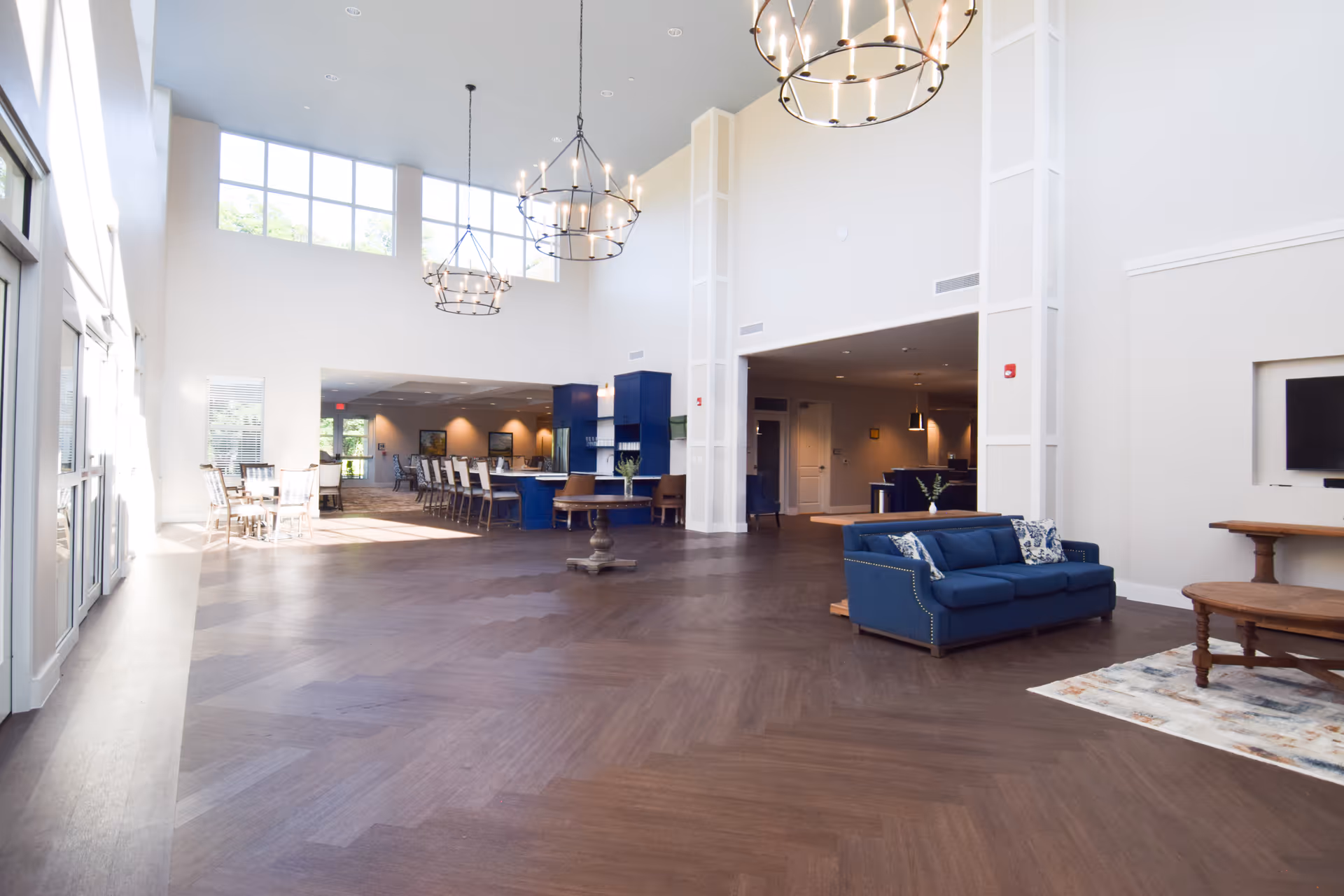 A spacious and bright common area in a senior living facility with high ceilings and large windows. The room features modern chandeliers, a blue sofa with patterned pillows, a wooden coffee table on a rug, and a dining area with tables and chairs in the background. The flooring is dark wood, and the walls are painted white, creating a clean and welcoming atmosphere.